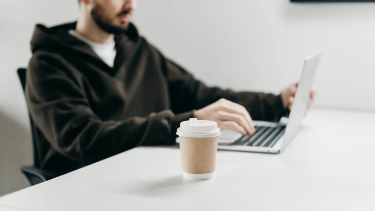 Productivity Hacks. A photo of a man at a white desk using his laptop, the man and laptop are out of focus and the camera is focusing on a cup of coffee.