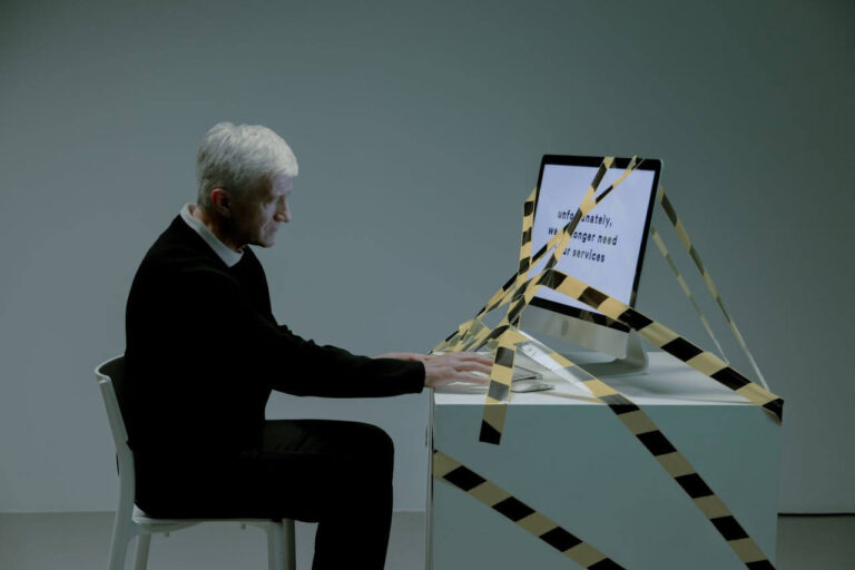 Redundant. A man sitting in an empty room witha computer that has 'your services are no loner required' on the screen, and yellow and black tape all on the desk and screen.